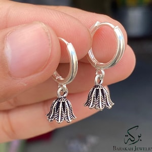 Puede incluir: Pendientes de aro plateados con colgantes en forma de campana. Los colgantes presentan un diseño detallado y texturizado con acentos oscuros. Los pendientes son sostenidos por una mano, mostrando el diseño de la joya. Barakah Jewelry.