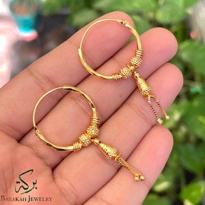 Puede incluir: Un par de pendientes de aro dorados con detalles intrincados. Cada pendiente presenta una serie de cuentas texturizadas y un dije colgante con delicados detalles de cadena. El nombre de la marca "Barakah Jewelry" es visible.
