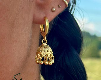 May include: Gold-colored hoop earrings with a bell-shaped charm. The charm features intricate filigree detailing and small dangling spheres. The earrings are worn on a person's ear, with the brand name "Barakah Jewelry" visible at the bottom.
