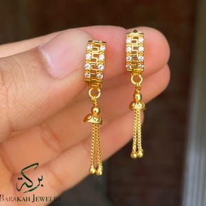 Op de afbeelding: Een paar goudkleurige oorbellen. De oorbellen hebben een rechthoekig bovendeel met heldere stenen en een bungelende ketting met kleine gouden kralen. De oorbellen worden in een hand gehouden, met de merknaam "Barakah Jewelry" zichtbaar.