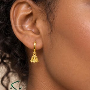 May include: A gold hoop earring featuring a small, dangling bell-shaped charm with textured vertical lines. The earring is secured through the earlobe, showcasing a polished metallic finish and a delicate, symmetrical design that hangs vertically.