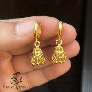 May include: A pair of gold-colored hoop earrings with a bell-shaped design. Each earring features intricate detailing and small dangling spheres at the bottom. The earrings are held in a hand, with the "Barakah Jewelry" logo visible.