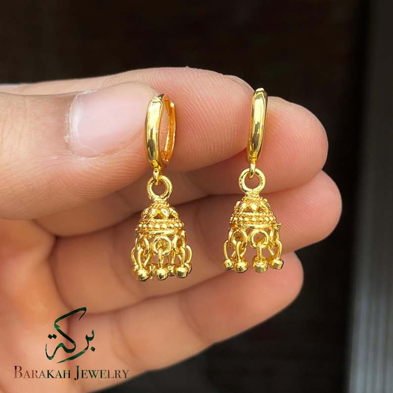 May include: A pair of gold-colored hoop earrings with a bell-shaped design. Each earring features intricate detailing and small dangling spheres at the bottom. The earrings are held in a hand, with the "Barakah Jewelry" logo visible.
