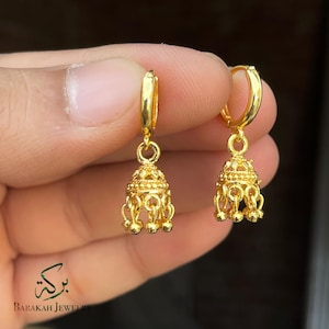 May include: A pair of gold-colored hoop earrings. Each earring features a bell-shaped charm with intricate detailing and small dangling accents. The earrings are held up against a blurred background.