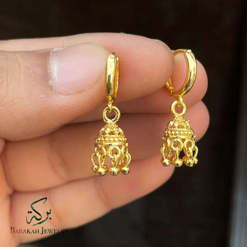 May include: A pair of gold-colored hoop earrings. Each earring features a bell-shaped charm with intricate detailing and small dangling accents. The earrings are held up against a blurred background.