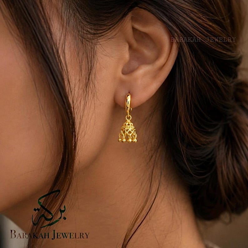 May include: A close-up of a gold-colored earring with a bell-shaped charm. The earring is a hoop style with a detailed, ornate design on the charm. The text "BARAKAH JEWELRY" is visible in the background.