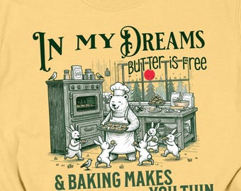 May include: Mustard yellow t-shirt with a whimsical illustration. The graphic features a bear chef, rabbits, and text that reads "In My Dreams Butter is Free & Baking Makes You Thin." The shirt has a crew neck and short sleeves.