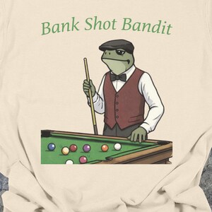 Könnte beinhalten: Elfenbeinfarbenes T-Shirt mit dem grünen Text "Bank Shot Bandit". Die Grafik zeigt einen Frosch in Weste und Mütze, der einen Billardqueue über einem Billardtisch mit bunten Kugeln hält. Das Shirt hat eine lockere Passform.