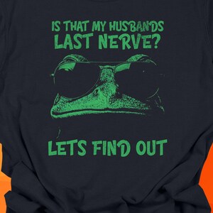 Green frog wearing sunglasses with the phrase “Is That My Husband’s Last Nerve? Let’s Find Out” in bold lettering. The humorous design pops sharply on the black Comfort Colors tee, giving it a playful, mischievous vibe perfect for Wives and women with sarcastic marriage humor.