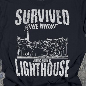 May include: A black short-sleeved t-shirt featuring a white graphic print. The design includes the text "SURVIVED THE NIGHT" above a lighthouse and trees, with the text "HUNTING ISLAND, SC LIGHTHOUSE" printed below the illustration.