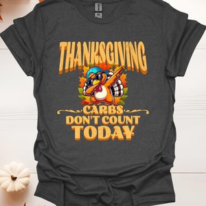 Puede incluir: Una camiseta gris oscuro con un gráfico de temática de Acción de Gracias. El gráfico presenta un pavo de dibujos animados con gafas de sol y una gorra de béisbol, haciendo el baile "dab". El texto en la camiseta dice "Thanksgiving Carbs Don't Count Today".