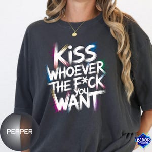 Puede incluir: Camiseta gris oscuro con el texto blanco "Kiss Whoever The F*ck You Want", con salpicaduras de pintura coloridas. La camiseta tiene cuello redondo y mangas cortas. La palabra "Pepper" está en un círculo.