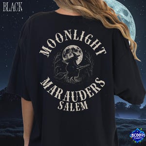 以下が含まれることがあります： 黒いTシャツ。背中には、満月の前をほうきに乗って飛ぶ魔女が描かれたグラフィックがあります。「MOONLIGHT MARAUDERS SALEM」の文字が画像の周りにアーチ状に配置されています。「BLACK」の文字が左上に印刷されています。