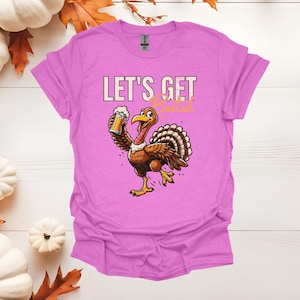 Puede incluir: Camiseta de color lila con un pavo de dibujos animados sosteniendo una jarra de cerveza. El pavo es marrón con una cola en abanico y patas amarillas. El texto "LET'S GET Basted" está impreso.