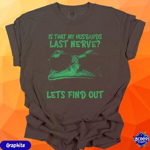 A vibrant green frog with sunglasses and the humorous text “Is That My Husband’s Last Nerve? Let’s Find Out.” The graphic stands out cleanly against graphite fabric, making it perfect for women who love sarcastic or mischievous humor.
