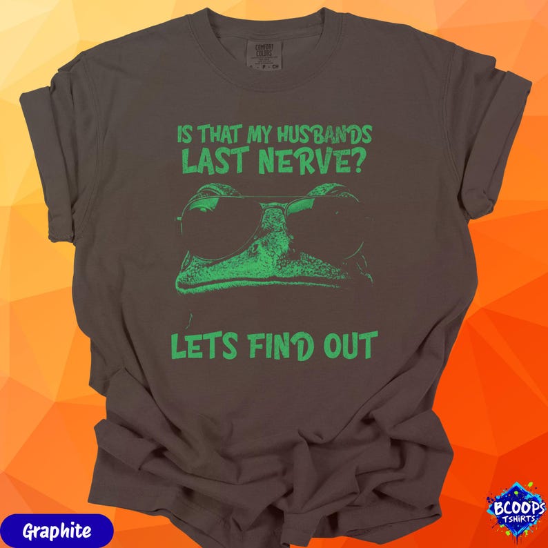 A vibrant green frog with sunglasses and the humorous text “Is That My Husband’s Last Nerve? Let’s Find Out.” The graphic stands out cleanly against graphite fabric, making it perfect for women who love sarcastic or mischievous humor.
