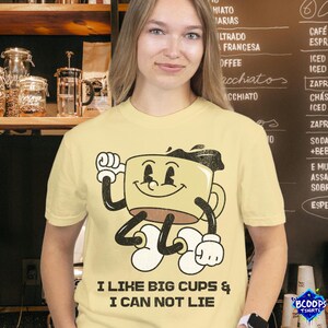 Puede incluir: Camiseta amarilla clara con un personaje de taza de café de dibujos animados. La taza tiene brazos y piernas negros, y guantes y zapatos blancos. El texto "I LIKE BIG CUPS & I CAN NOT LIE" está impreso debajo.