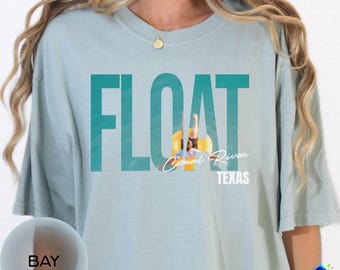 May include: A light blue t-shirt with the word "FLOAT" in teal block letters. The image within the "O" shows a person floating on a yellow tube in the Comal River, Texas. The shirt also has the words "Comal River" and "TEXAS".