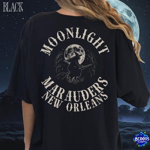 以下が含まれることがあります： 黒いTシャツ。背中には、満月の前をほうきに乗って飛ぶ魔女のグラフィックが描かれています。「MOONLIGHT MARAUDERS NEW ORLEANS」の文字が画像の上と下にアーチ状に配置されています。
