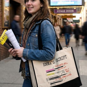 May include: A person stands on a city sidewalk holding playbills and a tote bag. The tote bag is cream-colored with black handles and features the text "EMMA'S SCRIPT" and other text. A Broadway theater marquee is visible in the background.