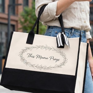 May include: A large tote bag with a cream-colored upper section and a black lower section. The bag features black handles and a black and white striped ribbon. The text "This Mama Prays" is printed in black within a laurel wreath design.
