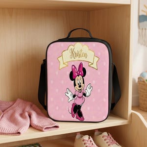 Puede incluir: Una bolsa de almuerzo rectangular con marco negro y panel frontal rosa con lunares blancos. La bolsa muestra un gr&aacute;fico de Minnie Mouse y una pancarta dorada con el nombre Kristen impreso en letras cursivas negras en la parte superior.