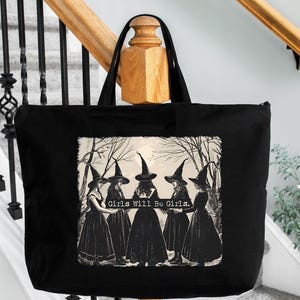 May include: A black tote bag with a white graphic of five witches in black cloaks and pointed hats. The witches are standing in a forest and the text 'Girls Will Be Girls.' is printed below them.