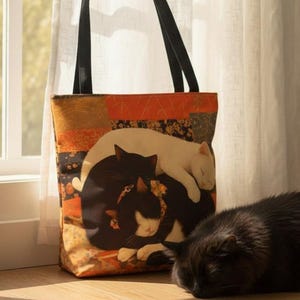 May include: A tote bag with a cat design and a sleeping black cat. The tote bag features a design of three cats, one white, one black, and one black and white, all curled up together. The bag has black handles.