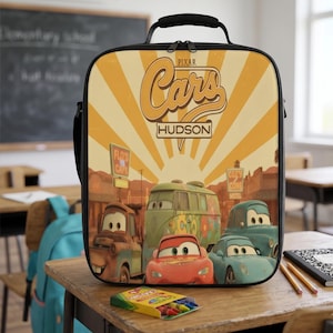 Puede incluir: Una bolsa de almuerzo rectangular con un gr&aacute;fico de Pixar Cars que muestra a los personajes Rayo McQueen, Mate, Fillmore y Sally. El dise&ntilde;o incluye un fondo de rayos de sol amarillos, el texto Cars y Hudson, y letreros de Flo's V8 Cafe y Cozy Cone.