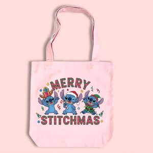 May include: A pale pink tote bag with a festive design. It features three blue cartoon characters and the words "MERRY STITCHMAS" in a plaid pattern, perfect for the Christmas season.
