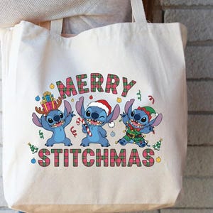 May include: A cream tote bag with a festive design. It features three blue cartoon characters and the text "MERRY STITCHMAS" in a plaid pattern. The characters are wearing Christmas-themed accessories.