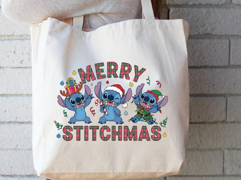May include: A cream tote bag with a festive design. It features three blue cartoon characters and the text "MERRY STITCHMAS" in a plaid pattern. The characters are wearing Christmas-themed accessories.