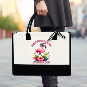 May include: A white tote bag with a black bottom and handles. The bag features a flamingo illustration wearing glasses and a hat, surrounded by flowers and leaves. Text on the bag reads "I DON'T GIVE A FLOCK I'M FABULOUS."