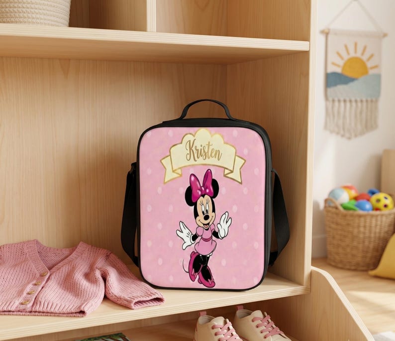 Puede incluir: Una bolsa de almuerzo rectangular con marco negro y panel frontal rosa con lunares blancos. La bolsa muestra un gr&aacute;fico de Minnie Mouse y una pancarta dorada con el nombre Kristen impreso en letras cursivas negras en la parte superior.