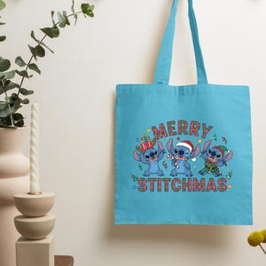 May include: A turquoise tote bag with a festive design. The bag features the text "MERRY STITCHMAS" above three blue cartoon characters in Christmas outfits. The bag has two handles and a simple design.