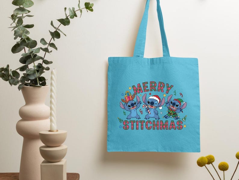 May include: A turquoise tote bag with a festive design. The bag features the text "MERRY STITCHMAS" above three blue cartoon characters in Christmas outfits. The bag has two handles and a simple design.