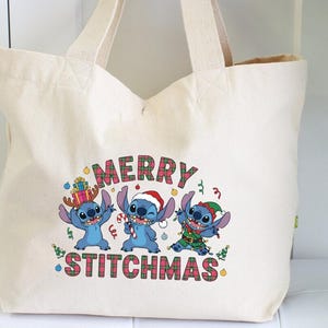 May include: Cream tote bag with a Christmas-themed graphic. The design features three blue cartoon characters in festive attire, with the words "MERRY STITCHMAS" in a plaid pattern.
