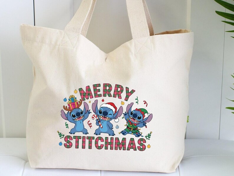 May include: Cream tote bag with a Christmas-themed graphic. The design features three blue cartoon characters in festive attire, with the words "MERRY STITCHMAS" in a plaid pattern.