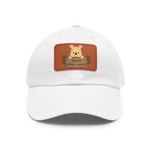 May include: White baseball cap with a brown leather patch featuring a cartoon Winnie the Pooh holding a sign that says "Hello Emma Macy".