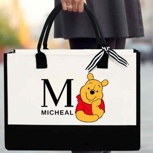 May include: White tote bag with black handles and a black bottom. The bag has a black and white ribbon bow and a Winnie the Pooh character with the letter "M" and the name "MICHEAL" printed on the front.