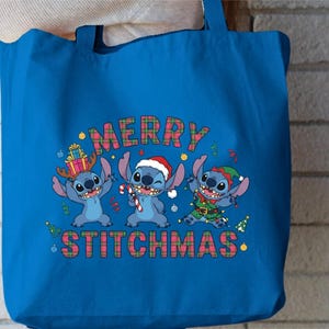 May include: A blue tote bag with a festive design featuring three cartoon characters. The text "MERRY STITCHMAS" is displayed in a plaid pattern. One character wears antlers, another a Santa hat, and the third an elf outfit.