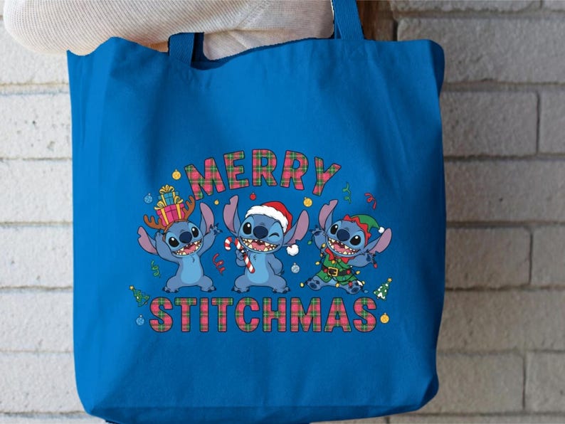 May include: A blue tote bag with a festive design featuring three cartoon characters. The text "MERRY STITCHMAS" is displayed in a plaid pattern. One character wears antlers, another a Santa hat, and the third an elf outfit.