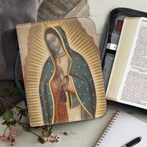 May include: A beige Bible cover featuring an image of Our Lady of Guadalupe with a gold halo and blue robe. The cover is next to an open Bible, a notebook, and a pen. The image has a vintage, religious theme.