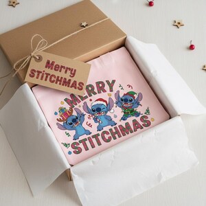 May include: A pink t-shirt with a festive design of three blue cartoon characters and the words "MERRY STITCHMAS". The shirt is presented in a brown gift box, accompanied by a tag that also reads "Merry Stitchmas".