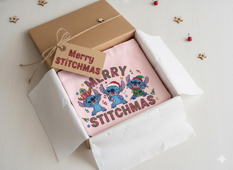 May include: A pink t-shirt with a festive design of three blue cartoon characters and the words "MERRY STITCHMAS". The shirt is presented in a brown gift box, accompanied by a tag that also reads "Merry Stitchmas".