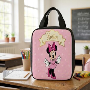 Puede incluir: Una bolsa de almuerzo rectangular con laterales y asas negras, que presenta un fondo rosa claro con lunares blancos. El frente muestra un gr&aacute;fico de Minnie Mouse y una pancarta dorada con el nombre Kristen impreso en letra cursiva.
