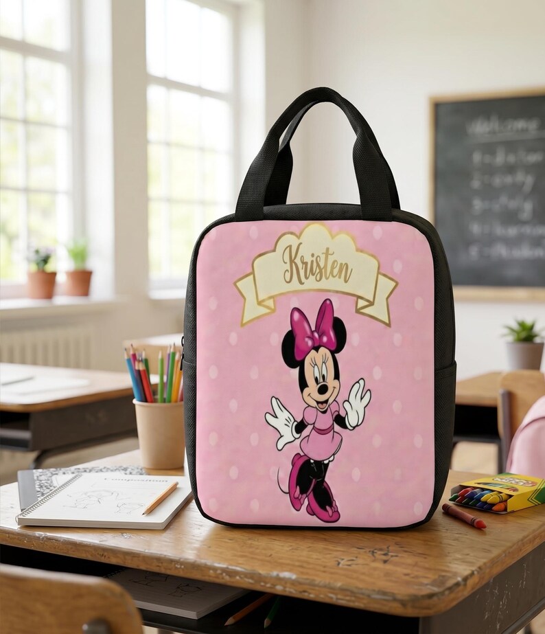 Puede incluir: Una bolsa de almuerzo rectangular con laterales y asas negras, que presenta un fondo rosa claro con lunares blancos. El frente muestra un gr&aacute;fico de Minnie Mouse y una pancarta dorada con el nombre Kristen impreso en letra cursiva.