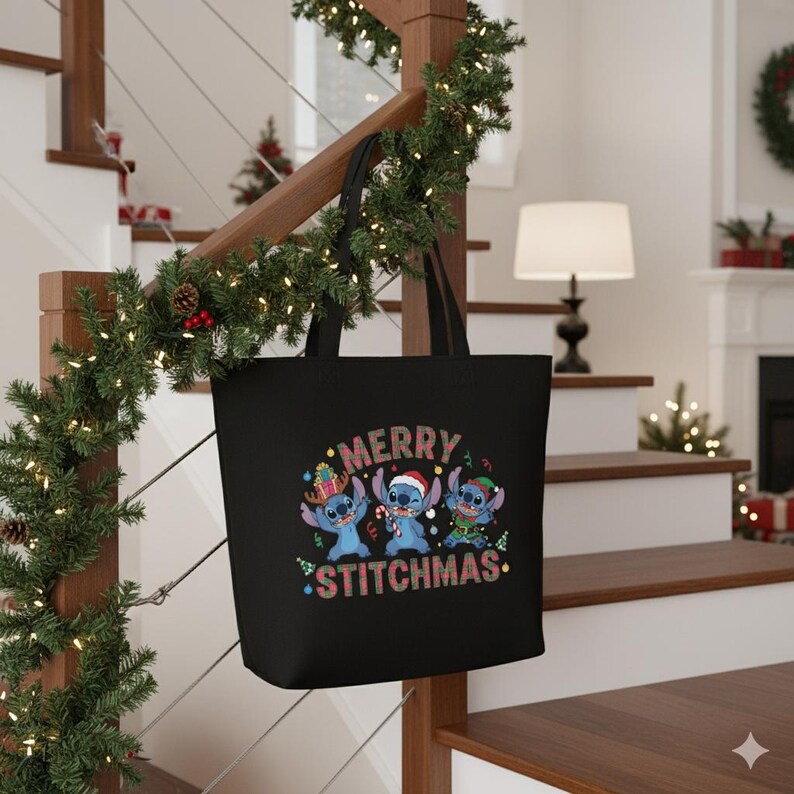 May include: Black tote bag with the text "MERRY STITCHMAS" and three blue cartoon characters wearing festive hats. The bag is hanging on a wooden staircase decorated with Christmas garland.