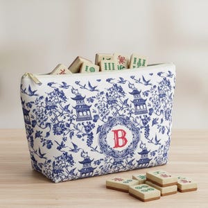 May include: A white zippered pouch with a blue floral and pagoda design, filled with mahjong tiles. The pouch features a red initial "B" in a decorative circle. Several mahjong tiles are scattered in front of the pouch.