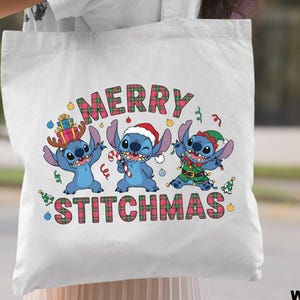 May include: White tote bag with a festive design featuring three blue cartoon characters dressed for Christmas. The words "MERRY STITCHMAS" are displayed above the characters in a plaid font. The bag is held by a person.